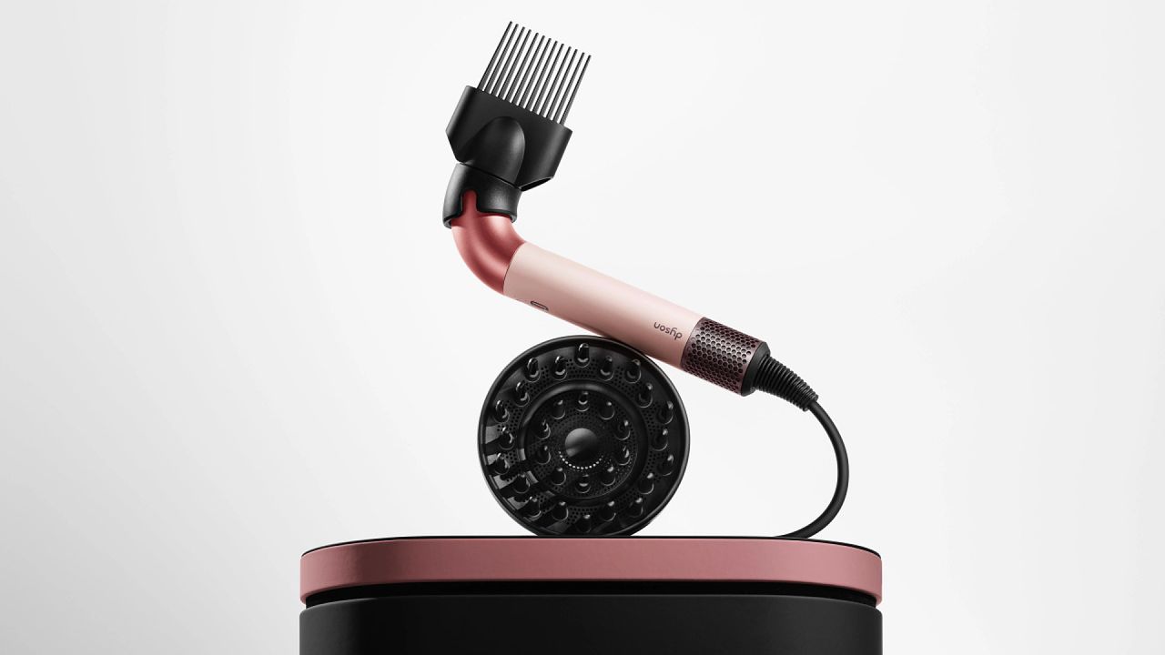 Dyson supersonic r blow dryer with wide-tooth and diffuser attachment sitting on pink and black carrying case against grey gradient background 