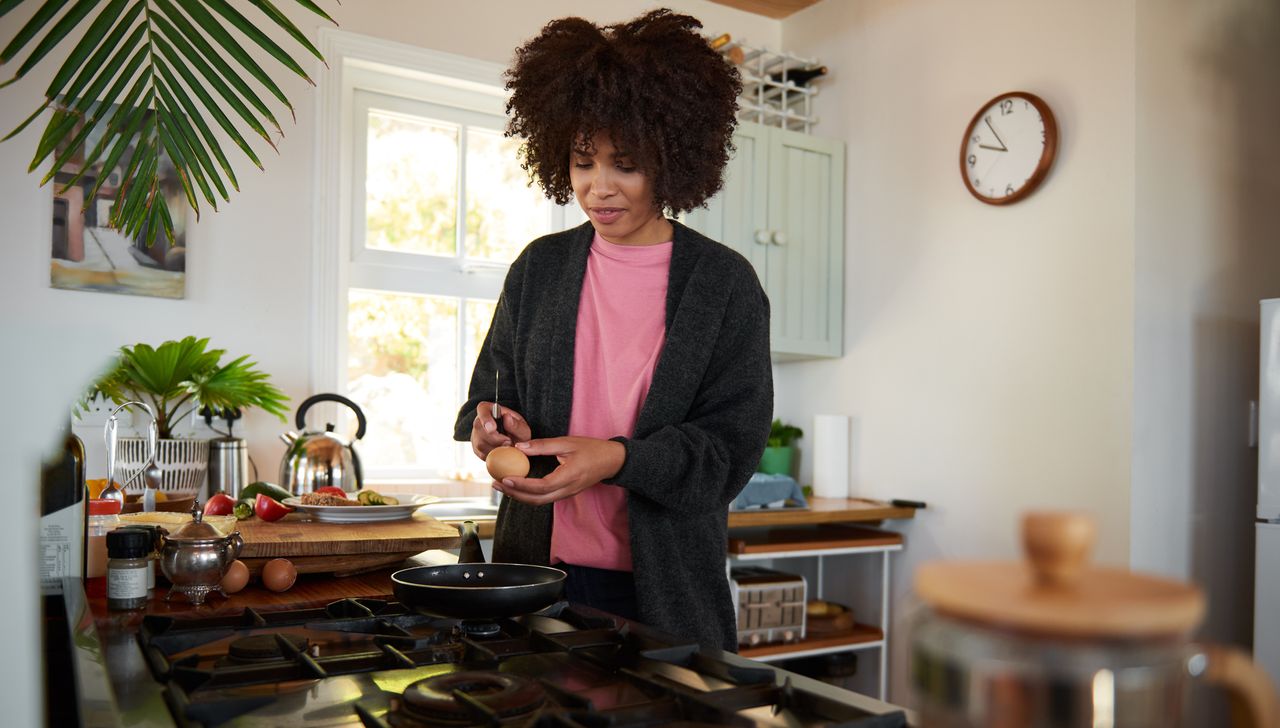 Woman in kitchen holding a knife and egg above a frying pan on a gas hob