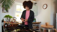 Woman in kitchen holding a knife and egg above a frying pan on a gas hob