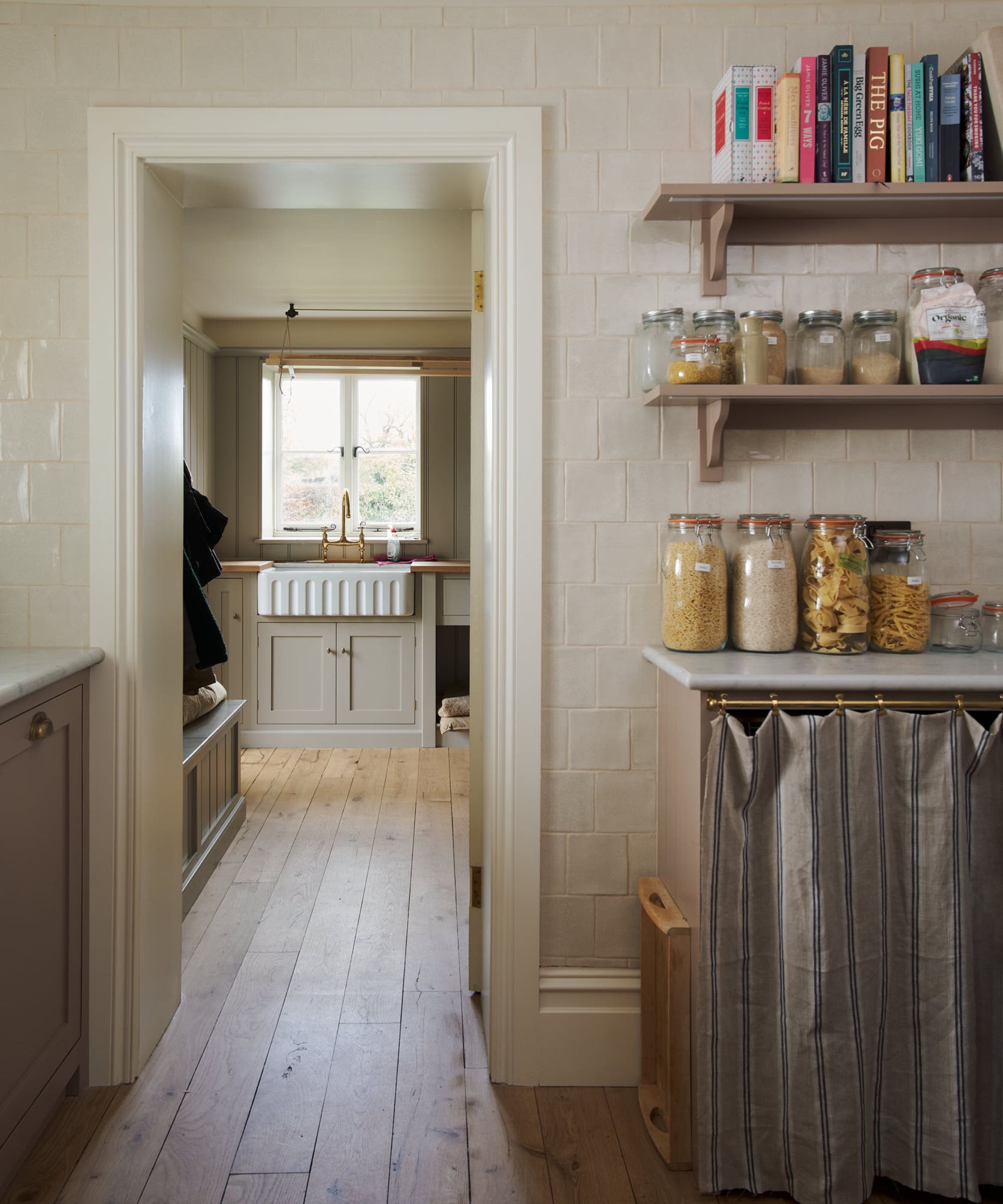 A country pantry and utility room with cream metro-tiled walls, open shelves stacked with glass storage jars, a Belfast sink with brass taps, painted cabinetry and wide plank timber floors.