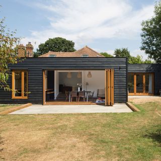 Black painted bungalow with open wooden stacker doors leading onto garden with tiled patio area in front