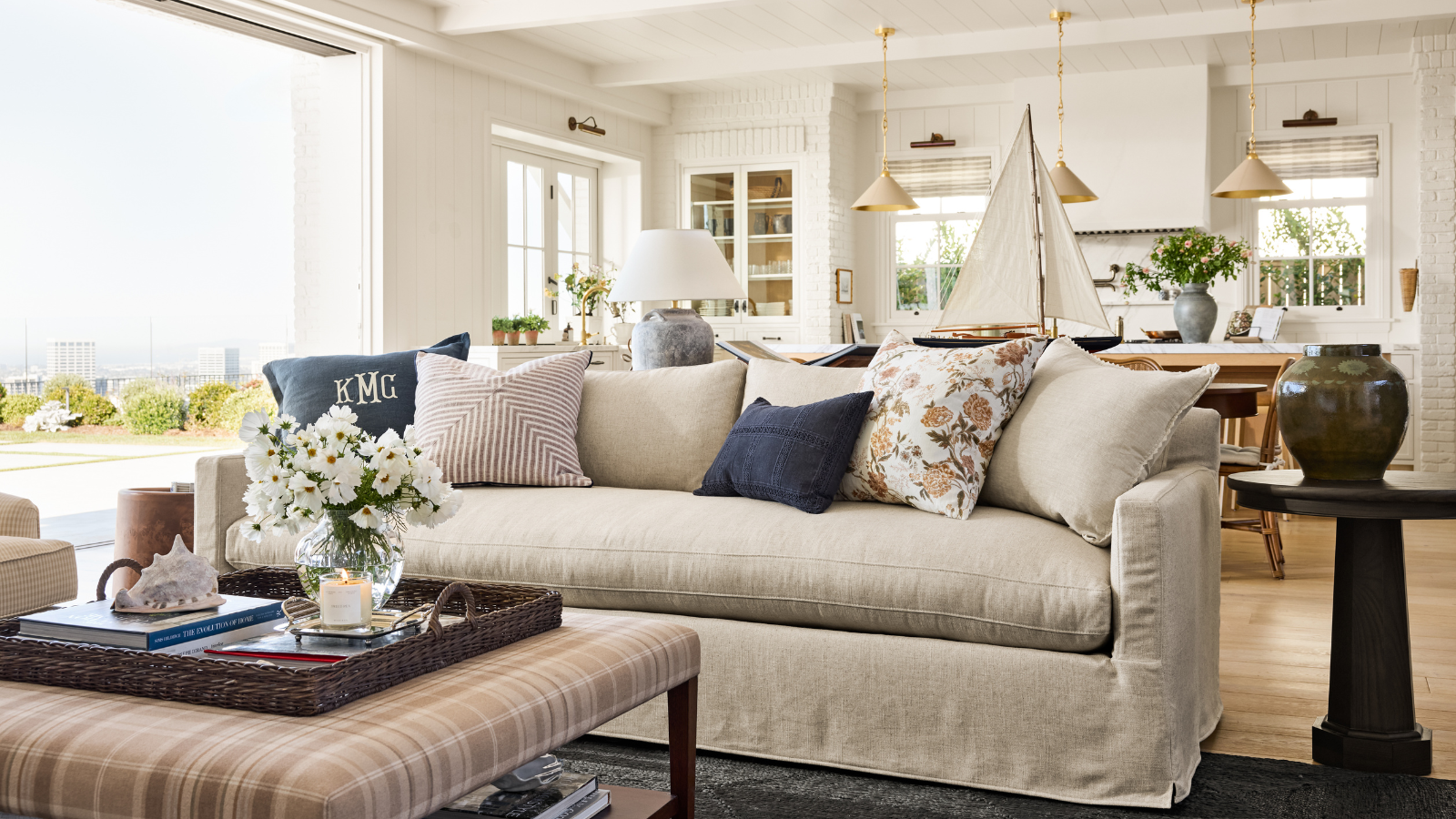a coastal-inspired living room with a large beige couch, several throw pillows, a plaid coffee table, decor on top, and a side table with a vase on top