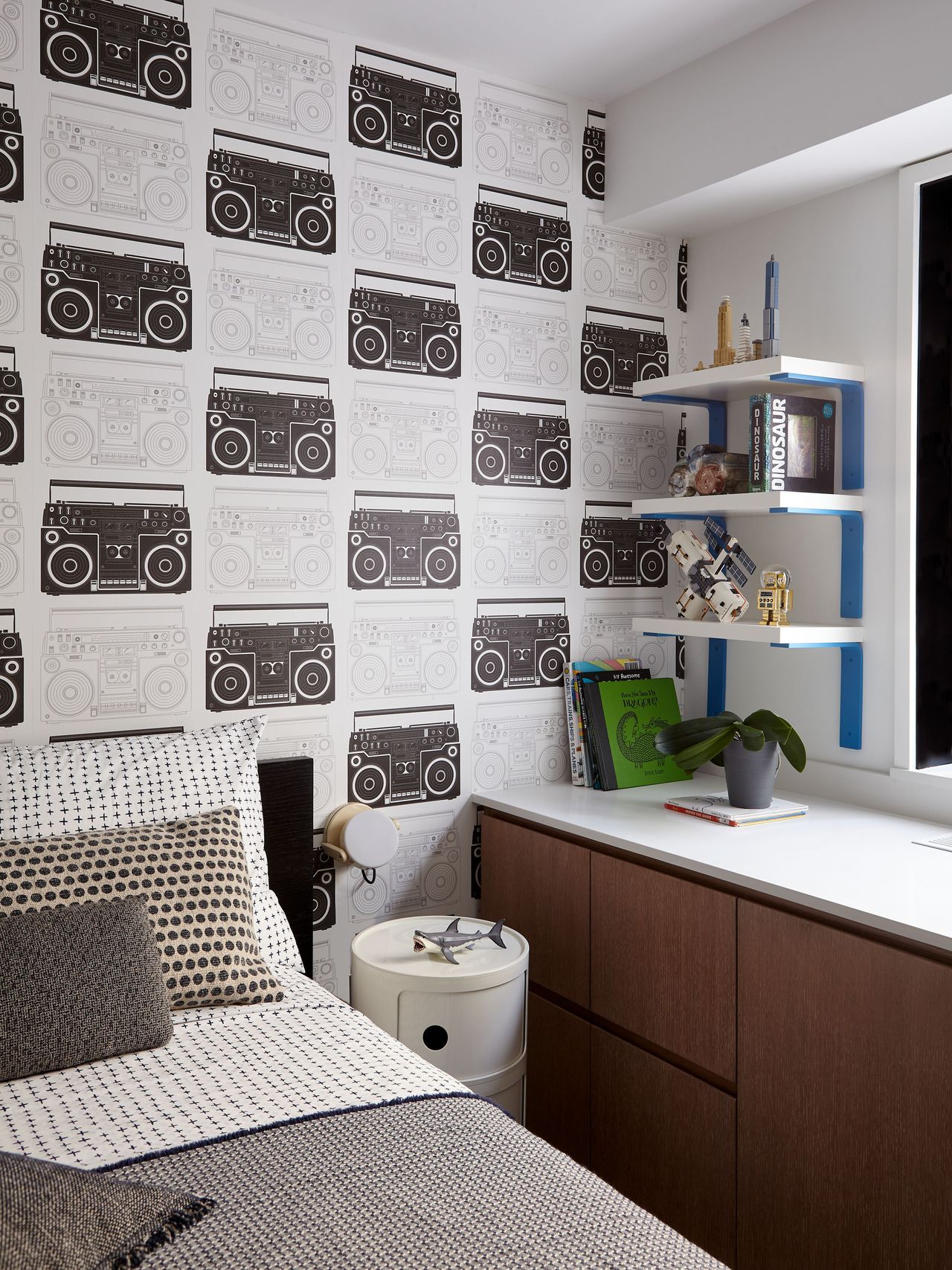a boys bedroom with black and white stereo design wallpaper and pattern clashing bedsheets in a predominantly gray hue with a desk area next to the bed with in-built storage and shelving above