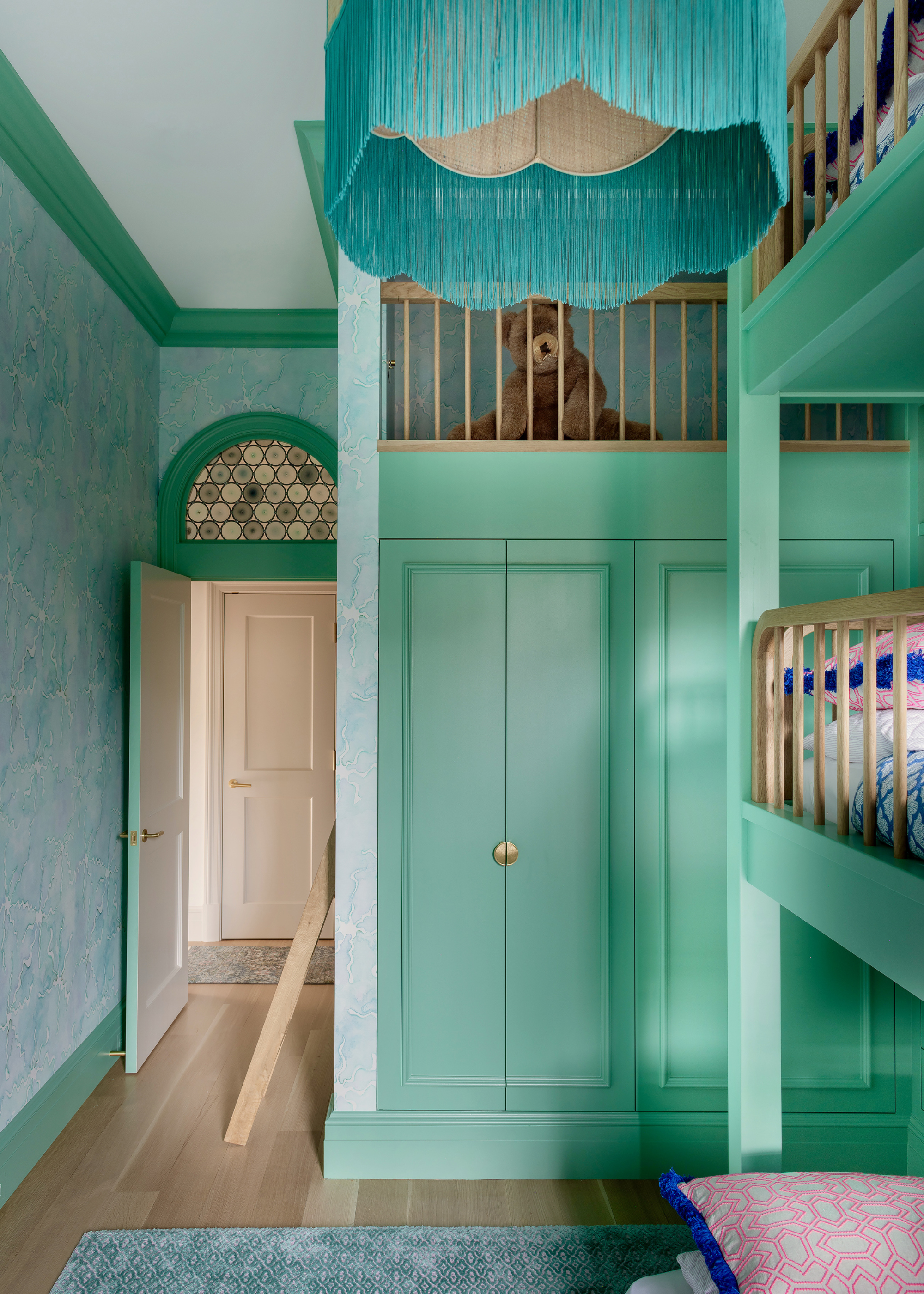 A children's bedroom with a three-tier bunk bed and built-in wardrobe painted in a vibrant green with a pale blue and green patterned wallpaper on the walls and an open doorway with an arched stained glass window above it
