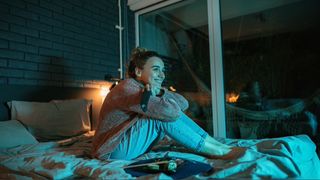 Woman sat in bed with gaming console in hand smiling at TV, lit by blue light.