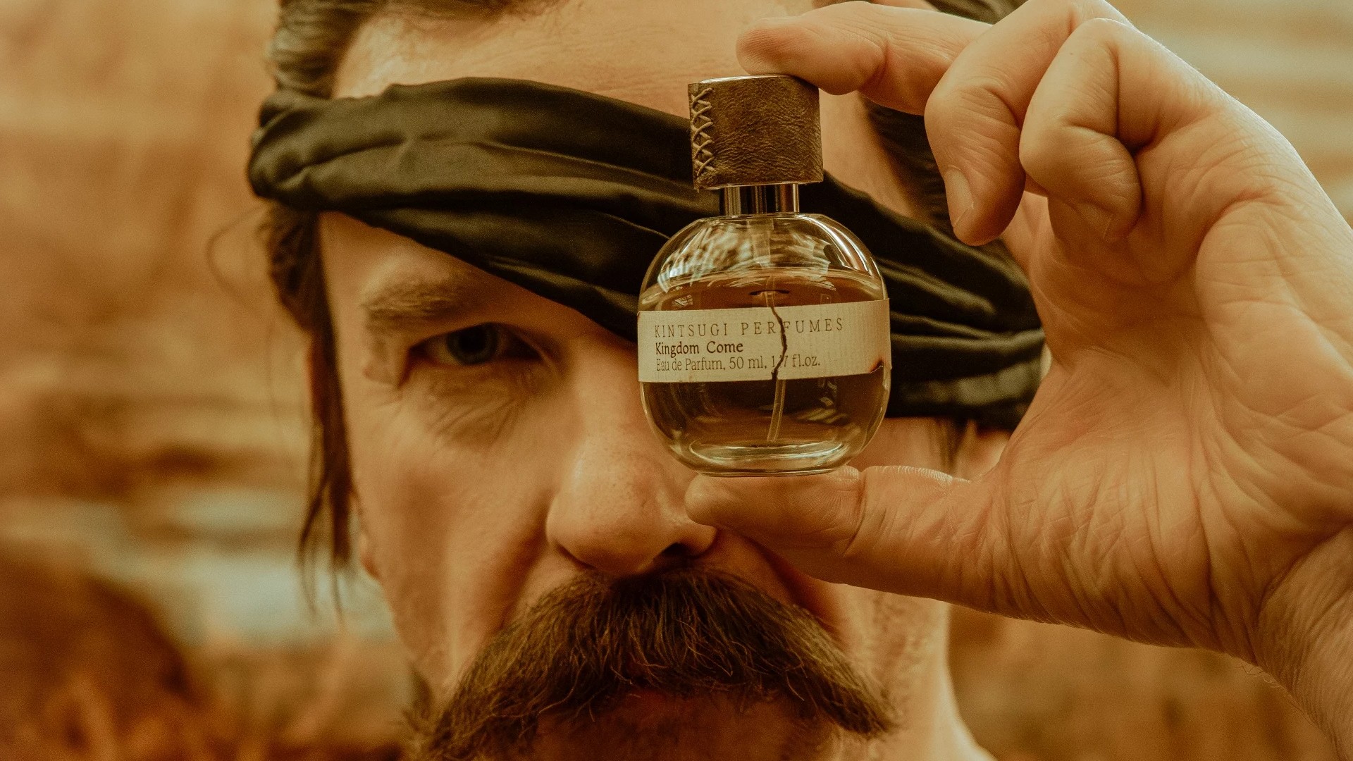 Man with one bandaged eye holds up bottle of Kingdom Come perfume.