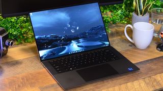 En Dell XPS 15 (2022) står öppnad på ett träfärgat skrivbord bredvid en vit kaffekopp. På skärmen visas en bakgrundsbild över ett landskap som skiftar i mörkblått.