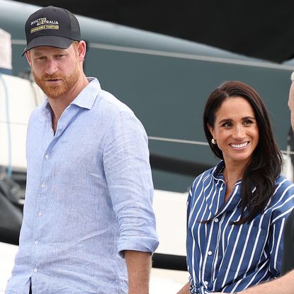 Prince Harry wears a baseball cap and a blue shirt, while Meghan Markle wears navy and white striped shirt and smiles