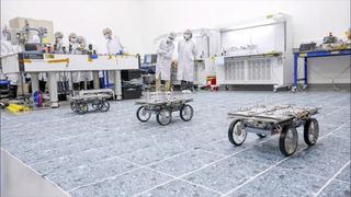 three small rovers with four wheels each pictured in a lab with scientists standing in the background to observe