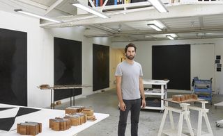 The artist Idris Khan in his studio with three large paintings on the walls with dark shapes on them.