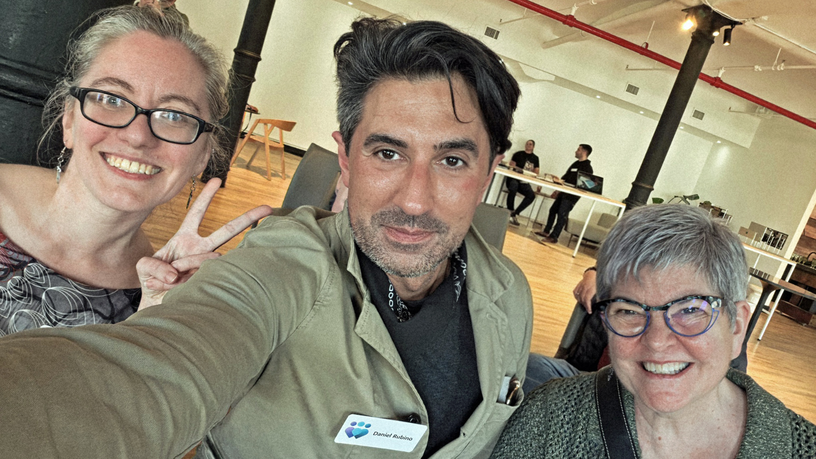 Three people smiling for a selfie in a well-lit room. The person on the left flashes a peace sign. The atmosphere is friendly and casual.