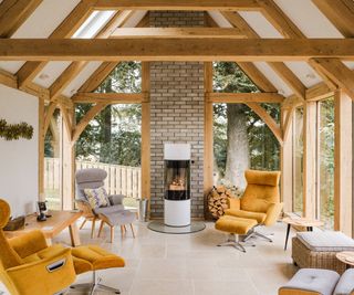 Glass and oak frame conservatory with yellow and grey armchairs positioned around a central freestanding modern woodburner