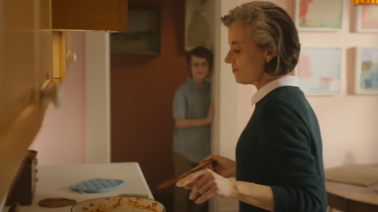 Chuck's grandmother dancing while watching a pot on the stove with a spoon in her right hand. Chuck is standing in the doorway behind her.