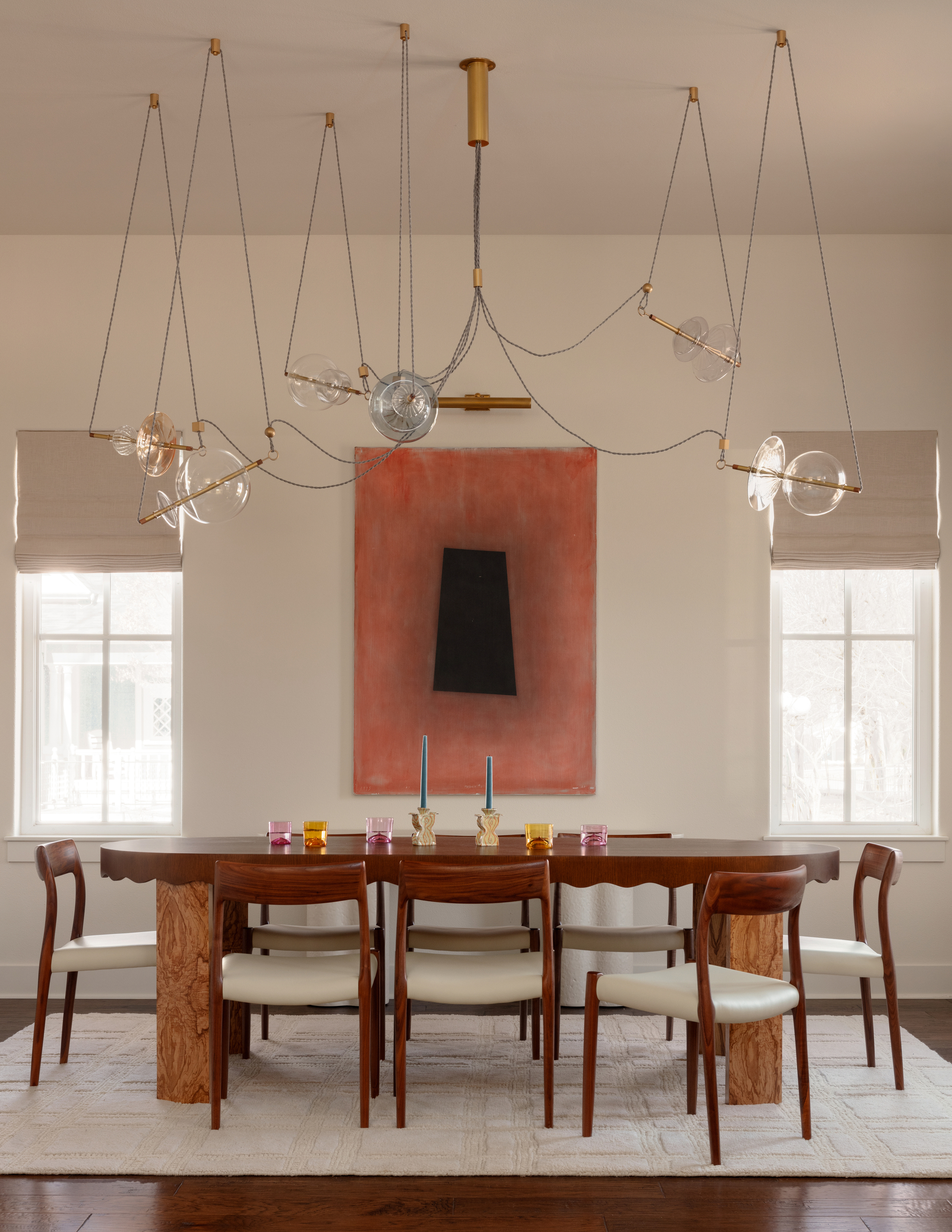formal dining room with timber table, upholstered dining chairs, large orange artwork, modern chandelier, colorful candle sticks on table, and two windows with roman blinds