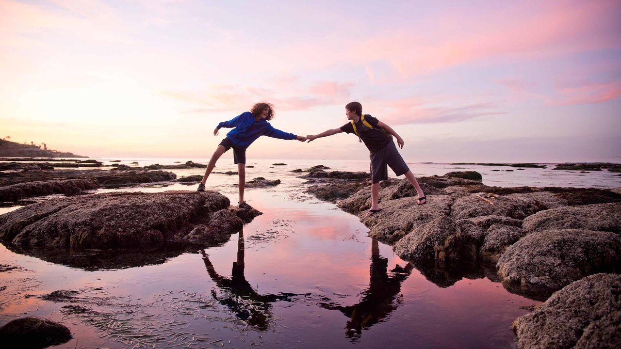 A woman reaches out her hand to help another to cross a stream.