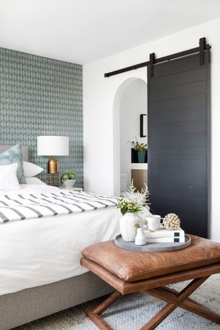 white bedroom with green graphic wallpaper, leather stool, sliding black door to ensuite, white bedding, copper side table lamp