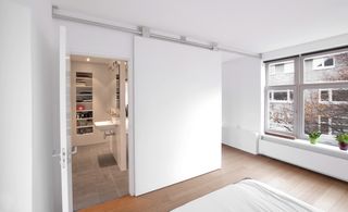 A room with white walls, ceilings and wooden floor. On the left is an open door that leads to the bathroom. On the right are clear glass window panels