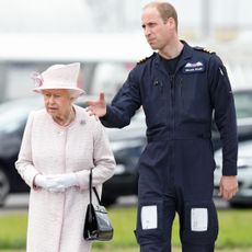Queen Elizabeth wears a light pink coat and matching hat and carries a black leather handbag, while her grandson Prince William wears his Royal Air Force uniform and guides her around