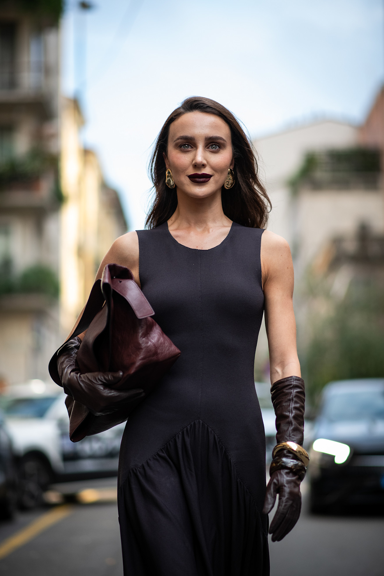 a woman with dark brunette hair and dark plum lipstick wears a dark purple dress with matching bag and leather gloves