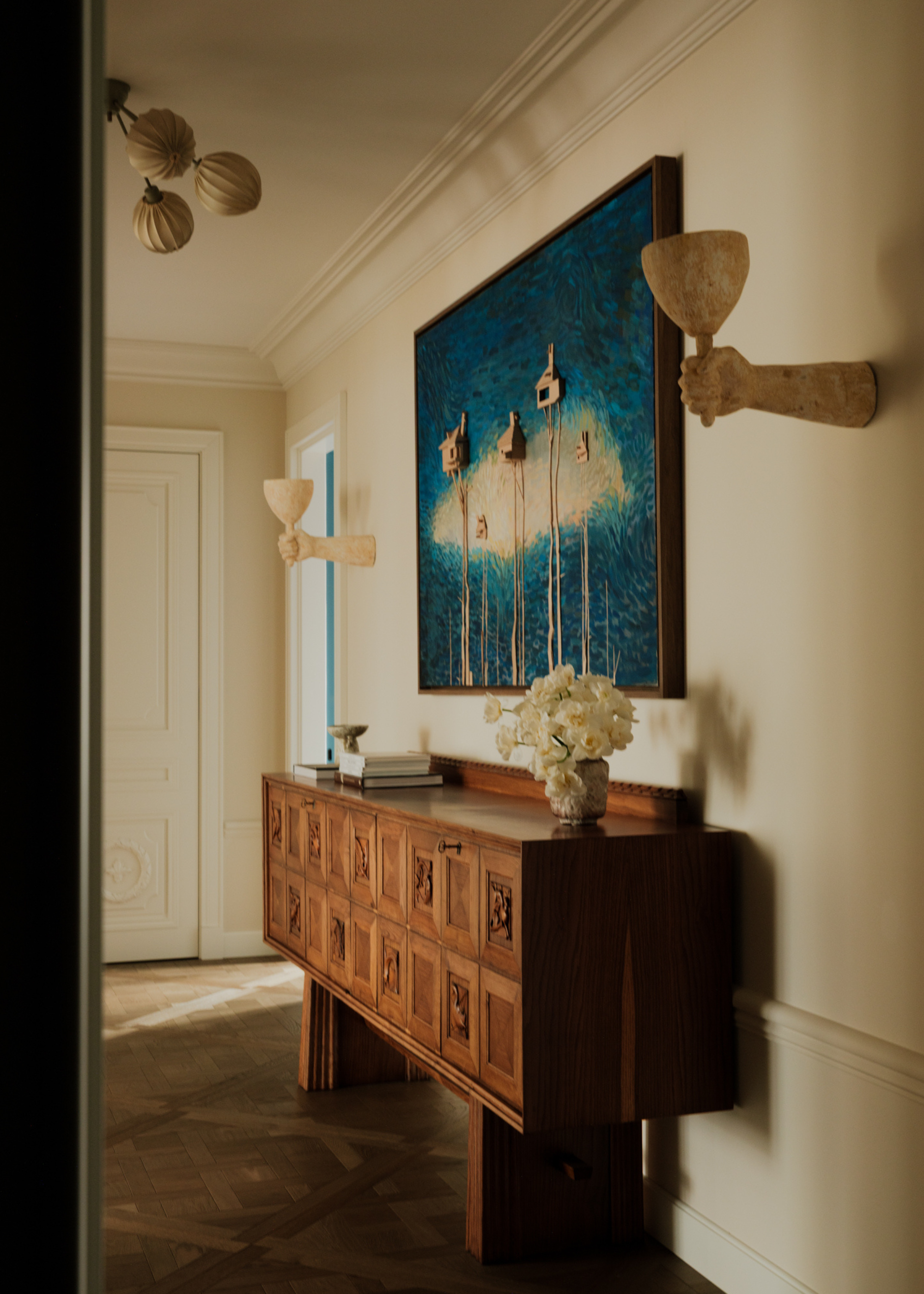 A white entryway with a wood sideboard, a framed blue painting, statuesque wall sconces, and a vase of flowers