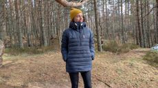 A woman in a forest wearing a blue down jacket and yellow hat with her hands in her pockets