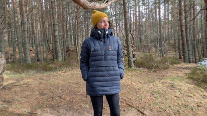 A woman in a forest wearing a blue down jacket and yellow hat with her hands in her pockets
