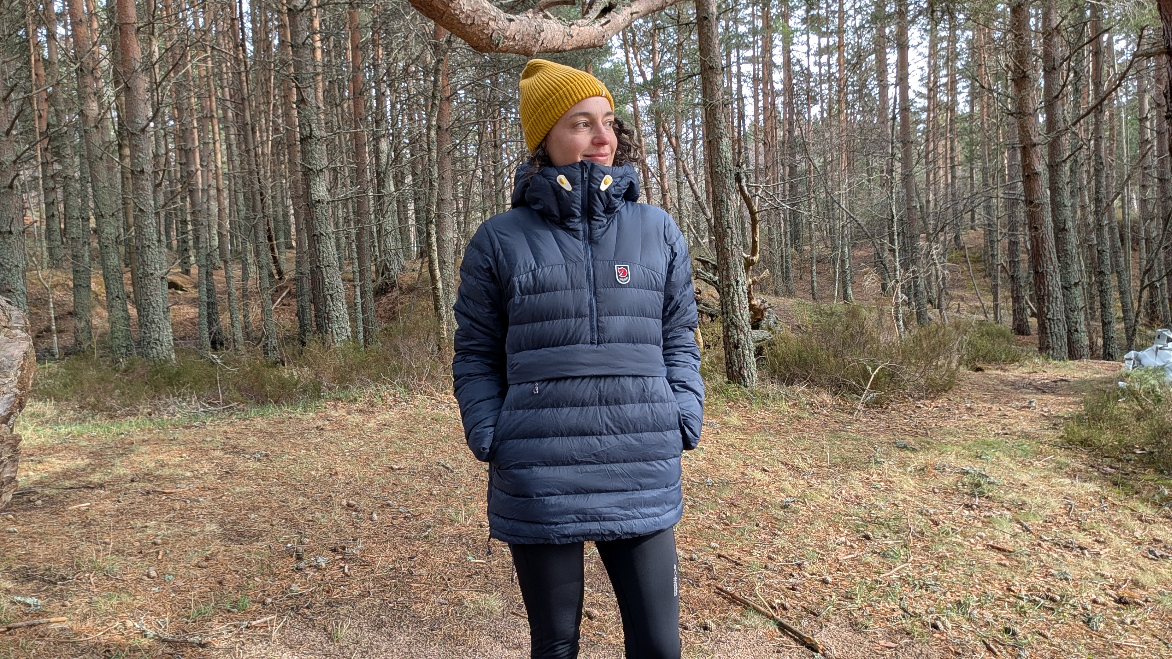 A woman in a forest wearing a blue down jacket and yellow hat with her hands in her pockets