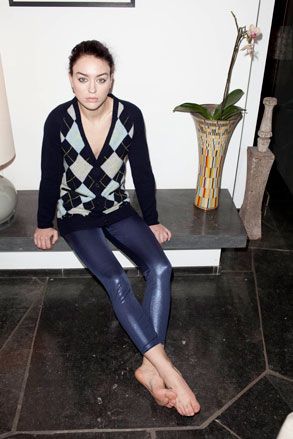 Female model sat on a slate grey bench,, wearing black and white diamond jumper, wet look blue leggings, bare feet, vase with tall flower, dark grey floor, white wall