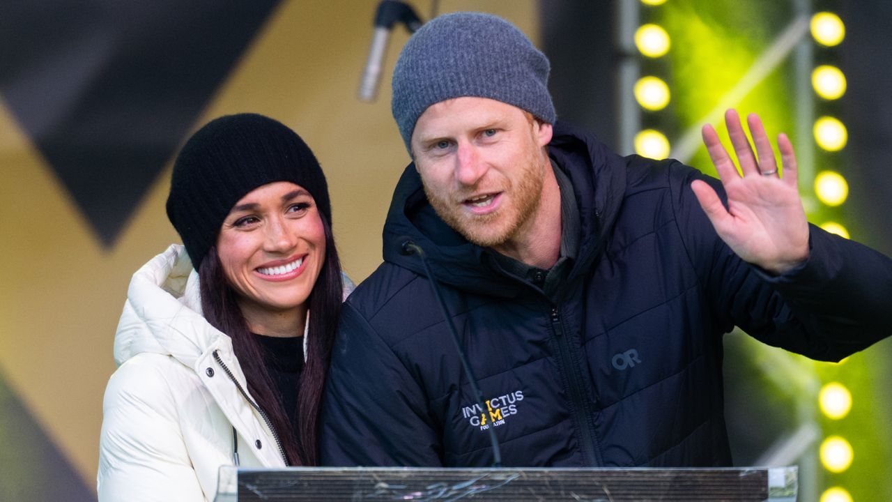 Meghan Markle wearing a white coat and black beanie smiling next to Prince Harry who is speaking at a podium wearing an Invictus Games jacket and waving