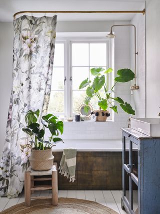 white bathroom with floral fabric shower curtain and shower over tub, blue vintage storage unit, plants, wooden floor, stool, round coir rug