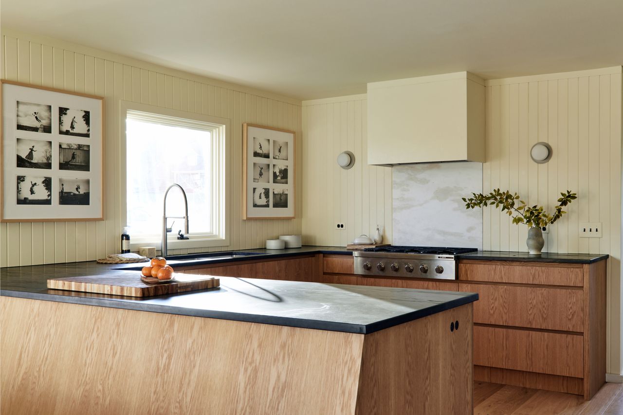 sustainable kitchen with wall panelling, sloped timber kitchen island and cabinets, dark stone counter, artwork hanging, and vase with flowers on bench