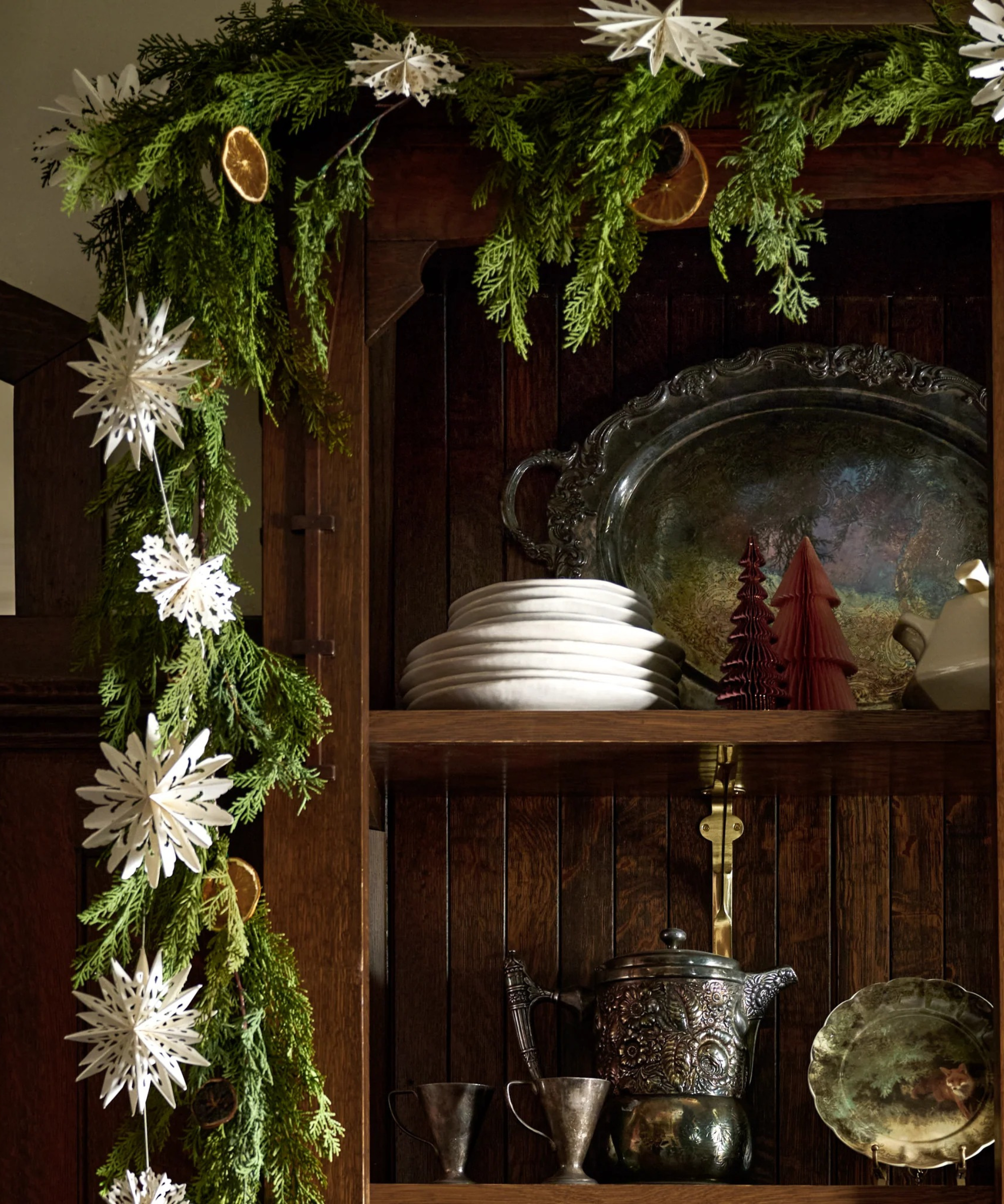a wooden cabinet with plates, a serving tray, and other kitchenware. Around the cabinet is garland and paper trimmings and dried fruit