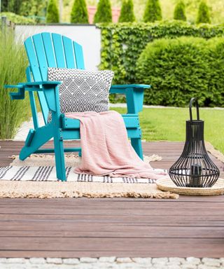 Blue Adirondack chair on a striped outdoor rug on a wooden patio with black bulbous lantern and green lawn border shrubs in background