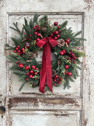 Red and green decorated Christmas wreath with cranberries and pine cones on it. The wreath is hanging on a rustic white wooden door