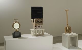 A display of items made from pebbles. On the left, is a medal-like sculpture with a black base and gold top. In the middle is 4 layers of bricks made out of pebbles with a black cement tray attached. On the right is a shovel with wood handles and blade made from gold pebbles.
