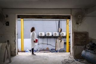 A young woman dressed in dark trousers and a white work shirt stands in the premises of an industrial workshop featuring cables and other utensils.