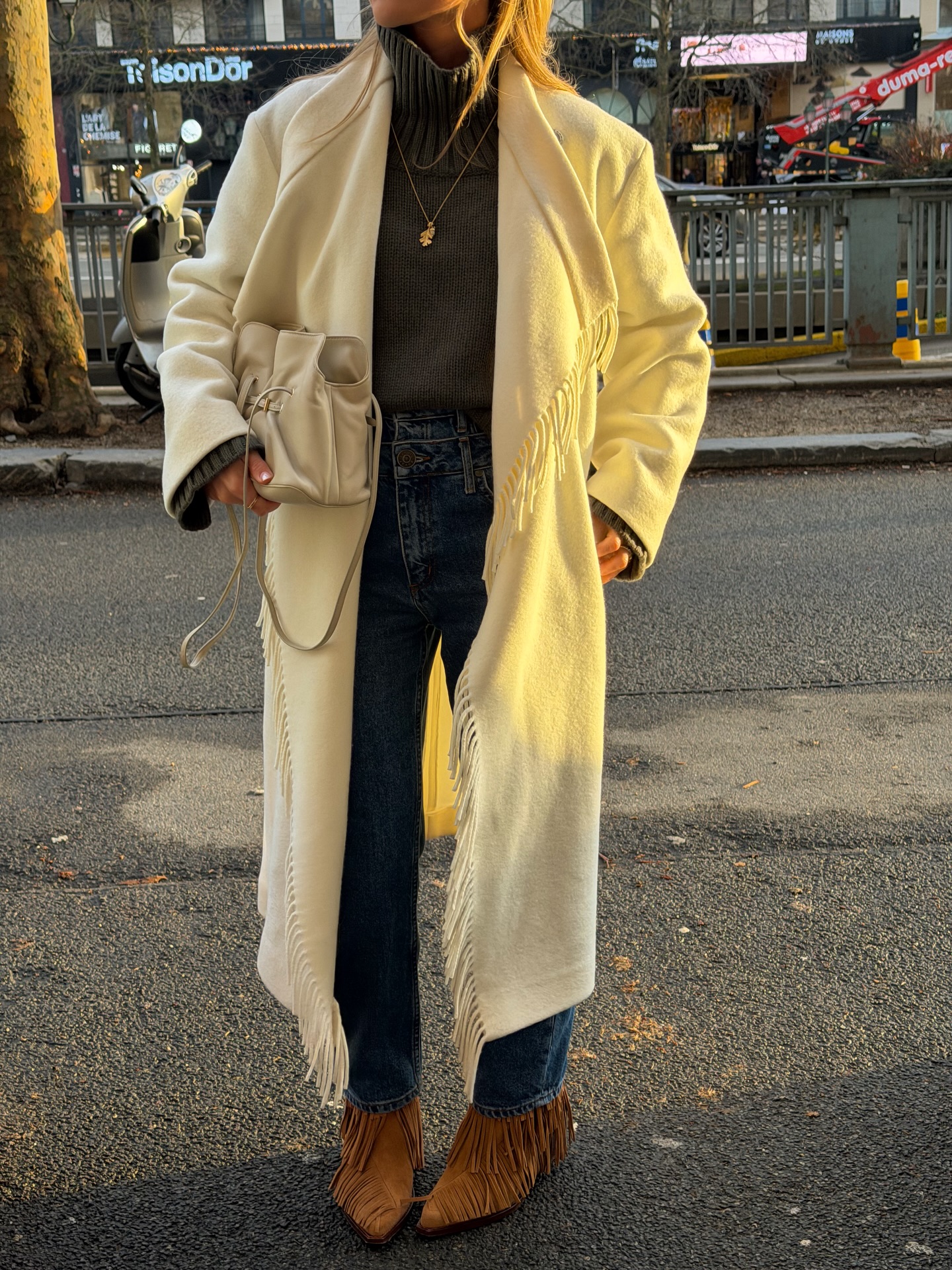 Fashion person Olivia Tps wears a Gen z spring outfit idea. She wears a fringed wool maxi coat roll neck jumper cigarette jeans and fringed boots in a recent image taken from her Instagram.