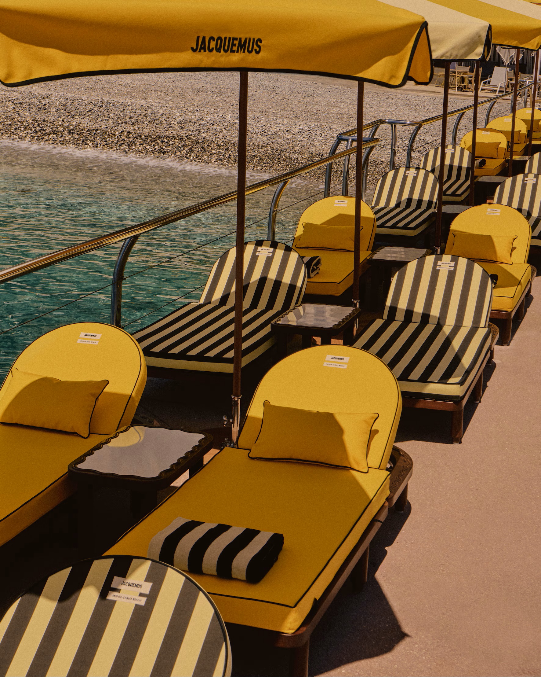 A series of yellow sun beds, some closer to ochre, some striped in pale yellow and black, is placed in two lines under matching parasols on a terrace overlooking the sea.