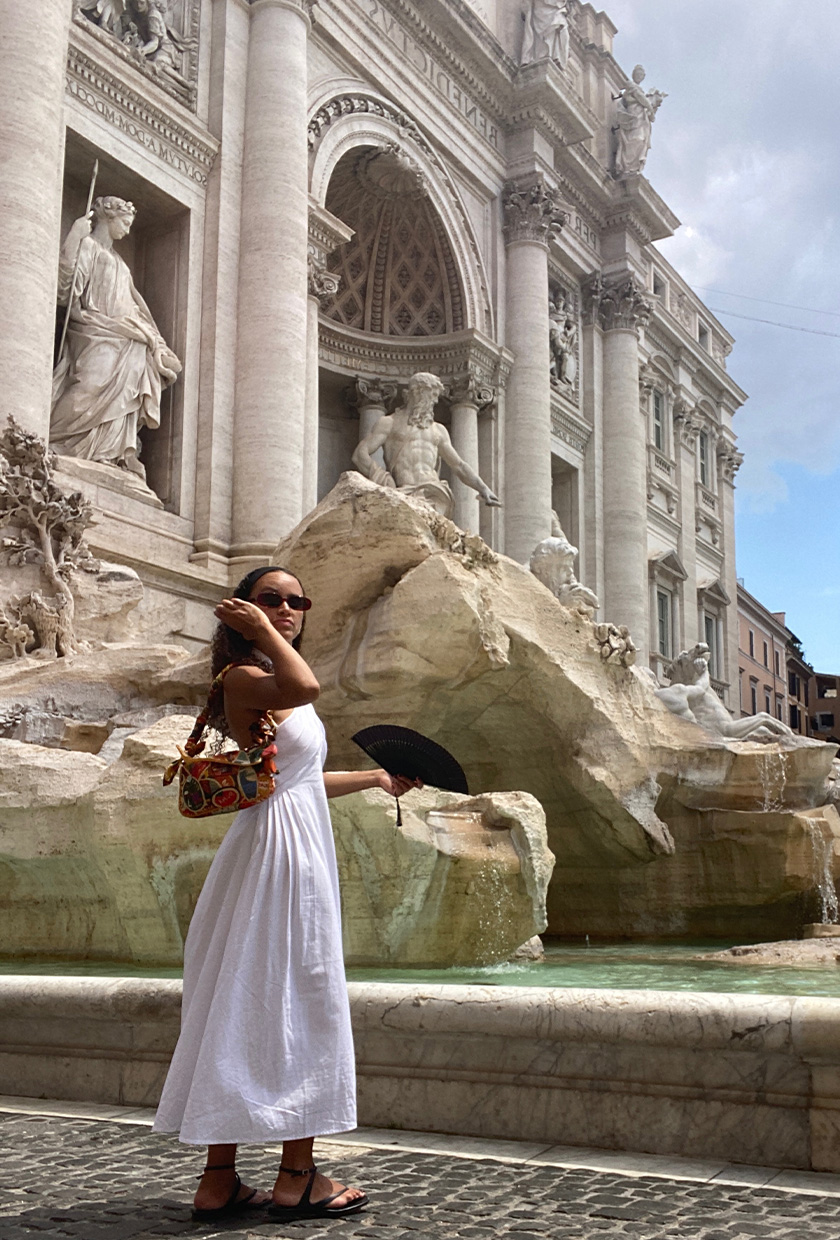 How to pack for Italy is shown in a picture of a fashion editor wearing a white linen dress, beaded shoulder bag, and black flat sandals