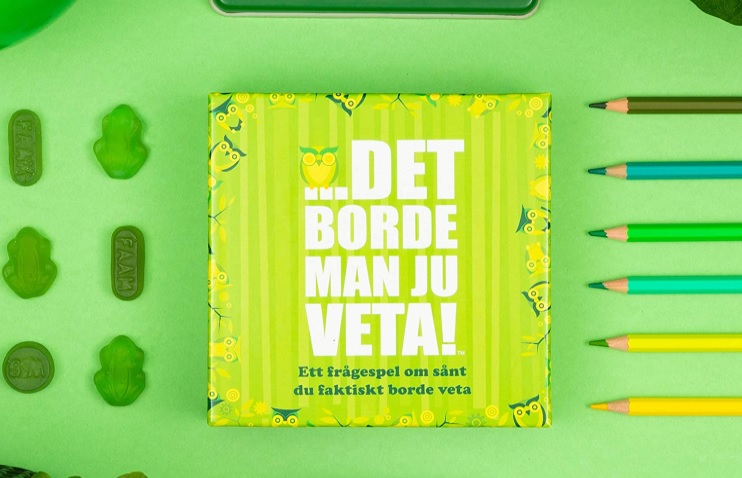 Sällskapsspelet Det borde man ju veta ligger på ett grönt bord bredvid ett par pennor och några gelégodisar.