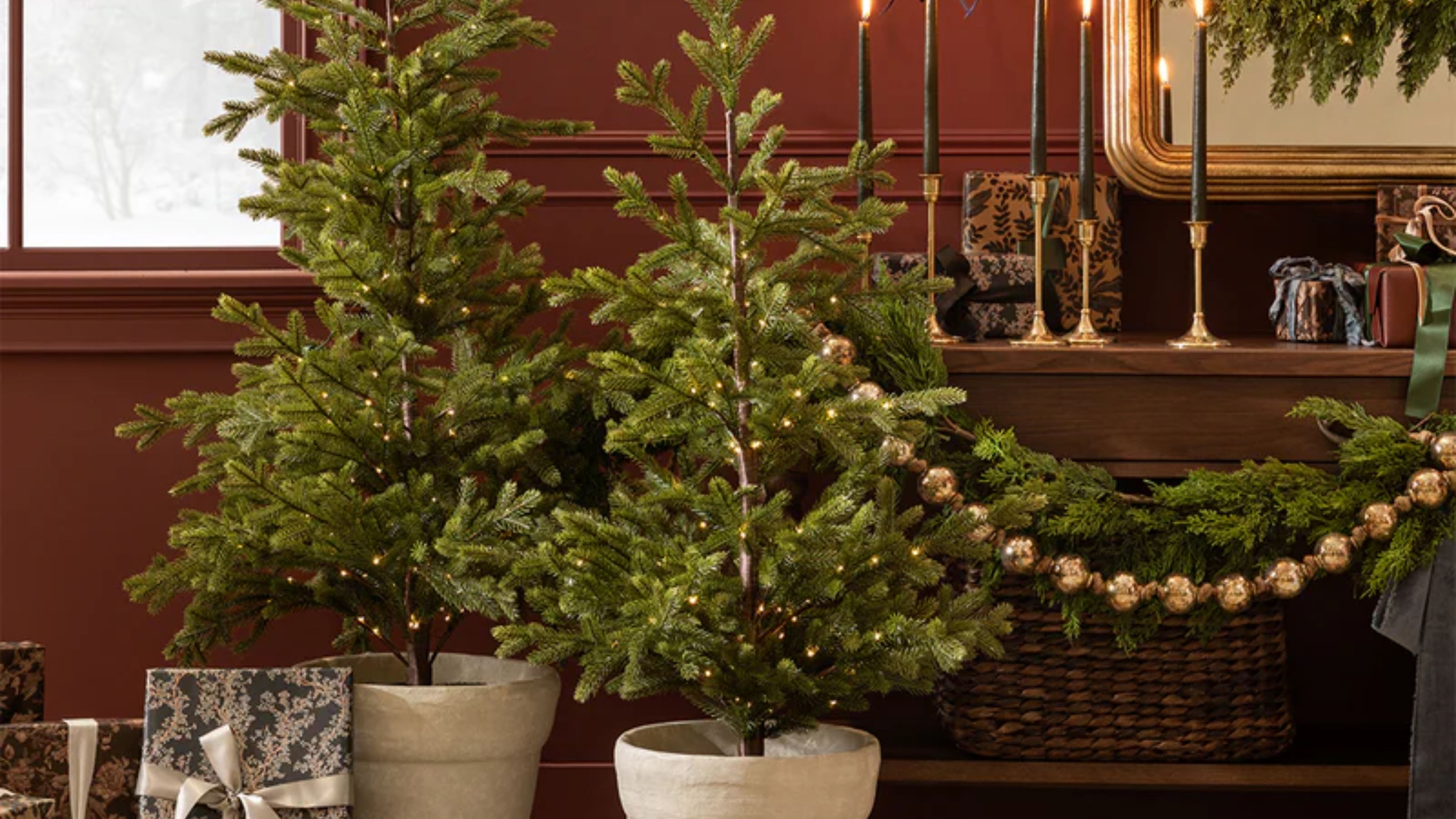 Mini pre-lit Christmas trees styled in a bordeaux living room area that features wrapped presents, golden candlesticks, and metallic beaded garland