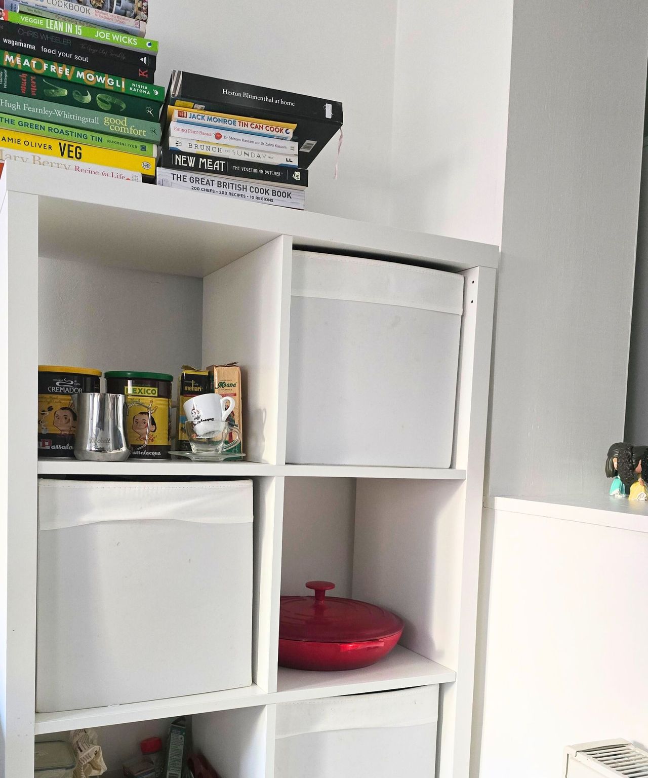 A white kitchen storage unit with colorful books on top of it and coffee accessories, a red kitchen pan, and white storage boxes in it