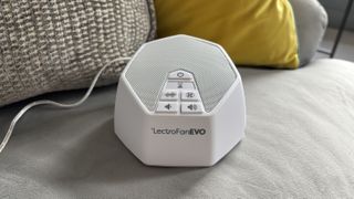 A white LectroFan EVO White Noise Machine perched on a grey couch with decorative cushions.