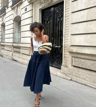 Influencer Ellie Delphine wears a white tank top, wicker basket bag, and dark denim maxi skirt while posing on Paris street.