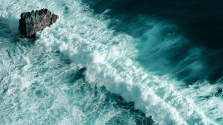 An aerial view of waves breaking on a rock and water foaming.
