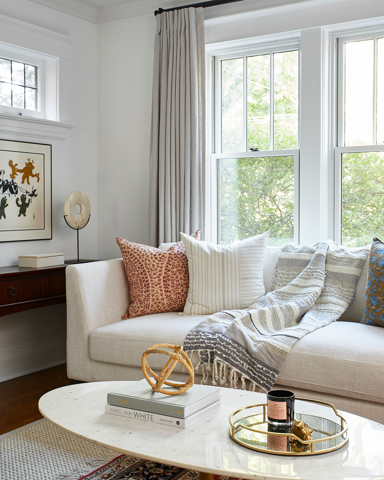 Image of the corner of a cozy white living room. There is a traditional white sofa with decorative throw pillows and a throw blanket. In front of the sofa is a white marble coffee table and there are windows behind the sofa.
