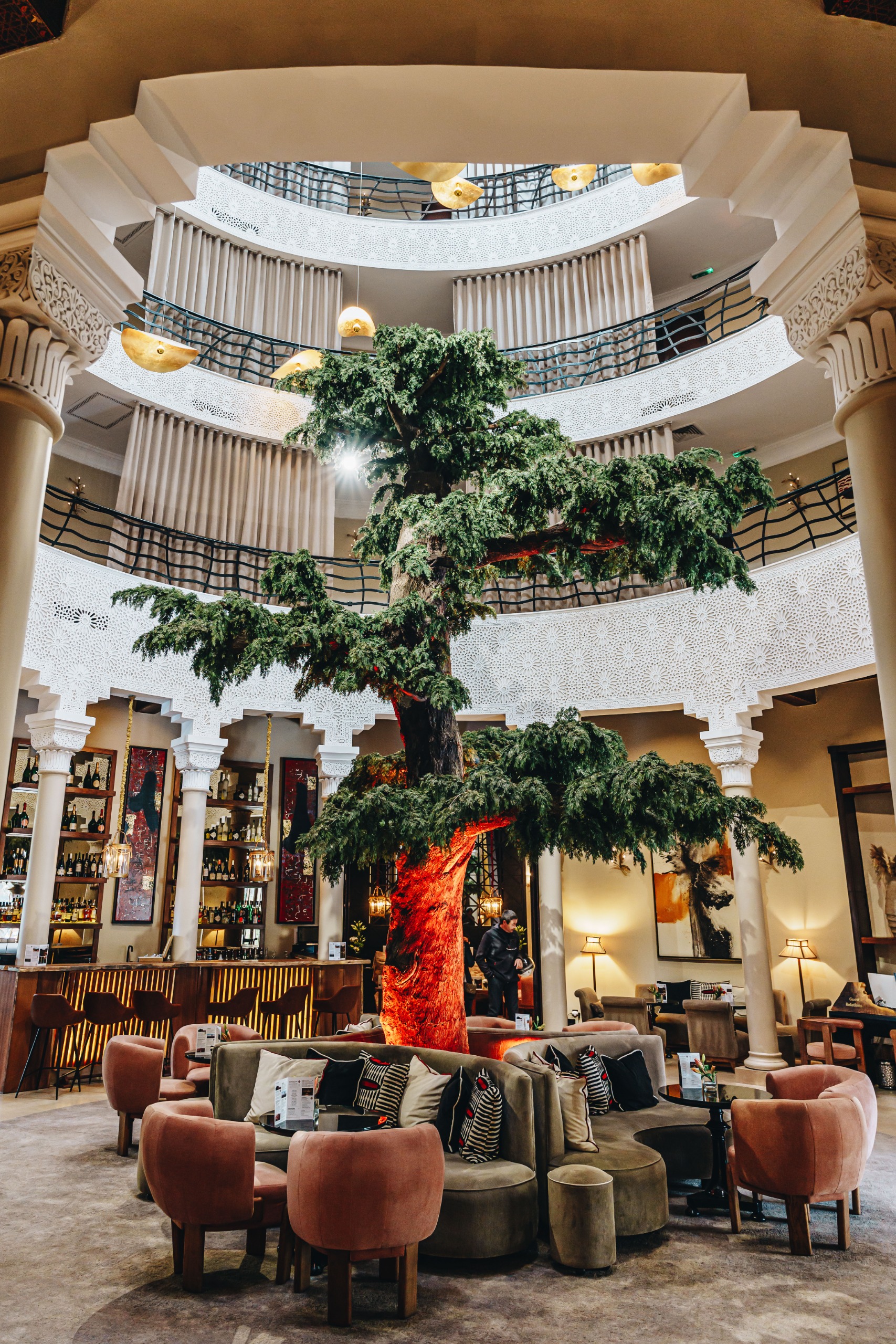 The plush fabrics-clad lobby of a Moroccan hotel with a central tree around which are placed a series of dark green and pink banquettes and chairs and golden pendant lights.