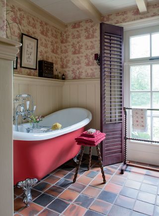 cast iron slipper bath with red sides with part paneled walls and pink toile de Jouy wallpaper window shutters and vintage accessories