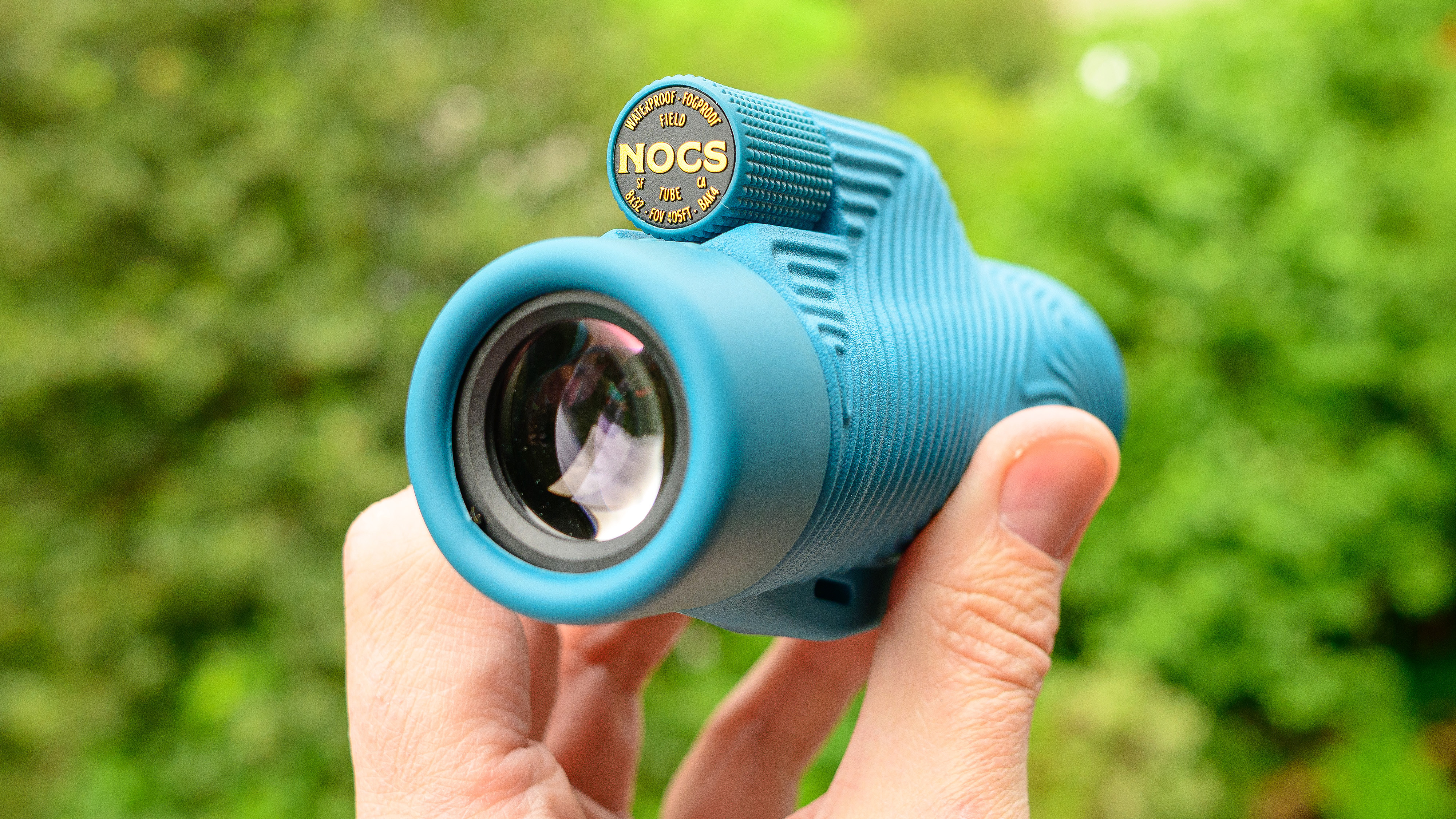 Nocs Field Tube Monocular in baby blue. Shown in a user's hand against a green background.