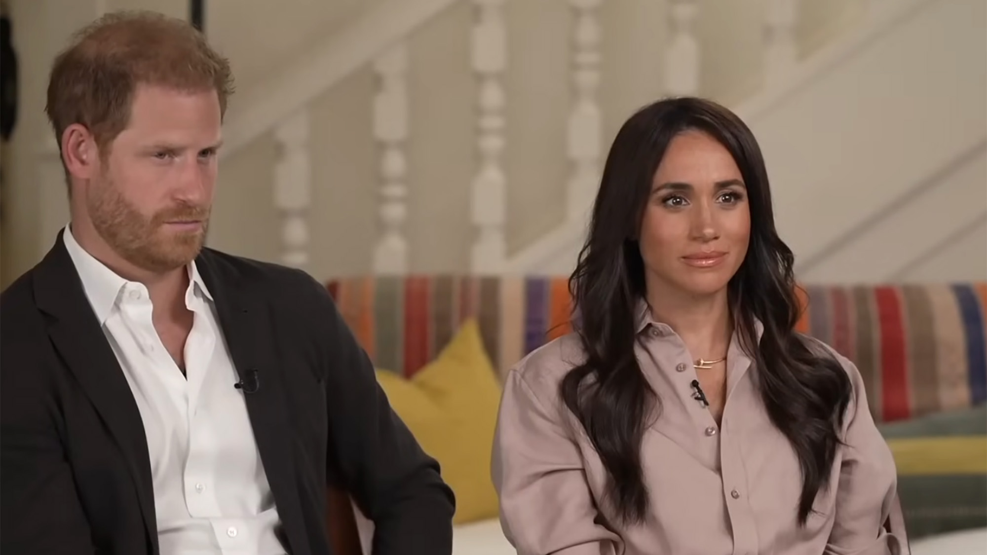 Prince Harry and Meghan Markle (Sussex) sit down for an interview in dress clothes. They are set up with a stairwell in the backdrop.
